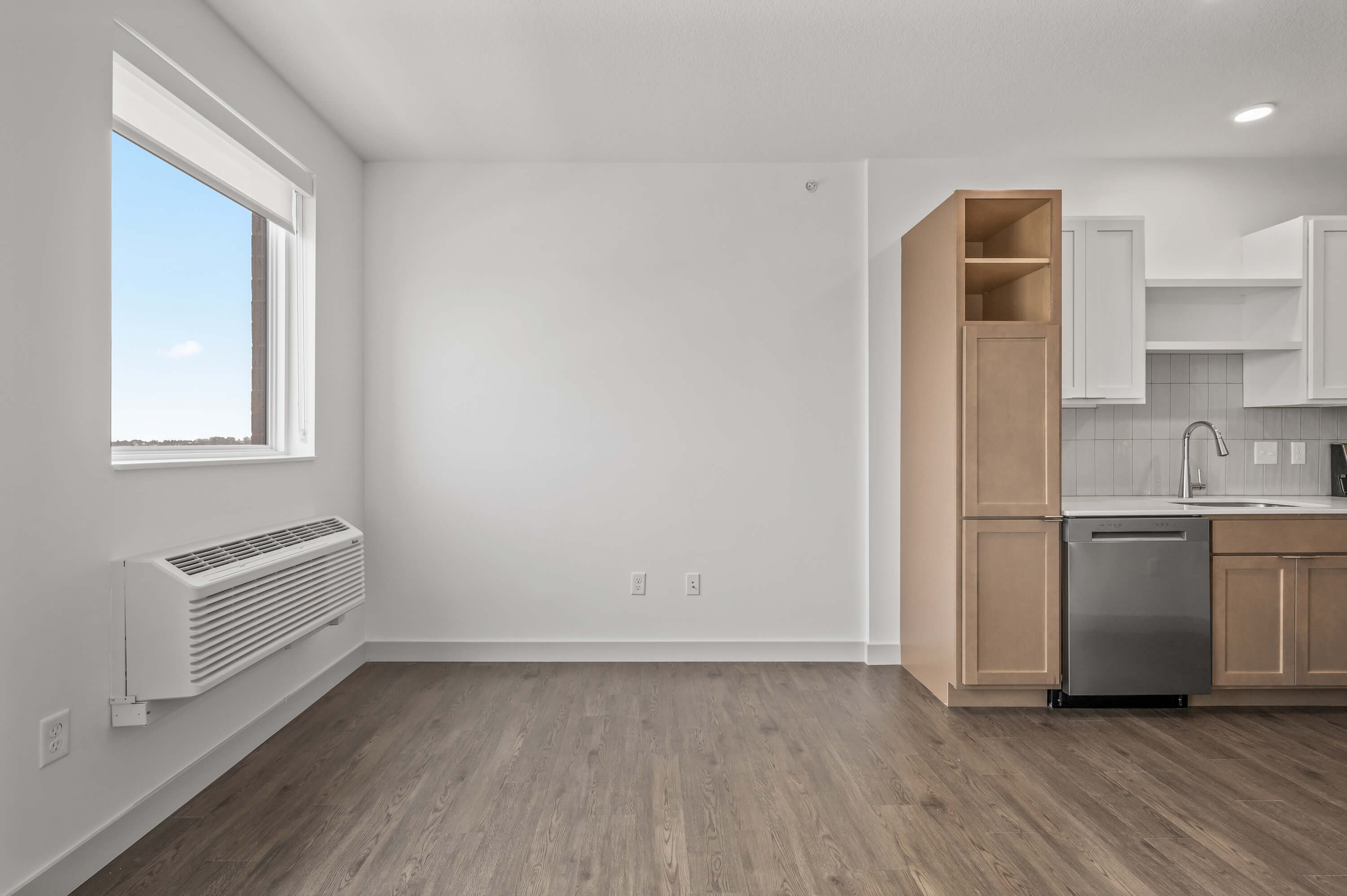 Corner of the living room with a kitchen in the background, showing cabinetry, a dishwasher, and a wall-mounted air conditioning unit.