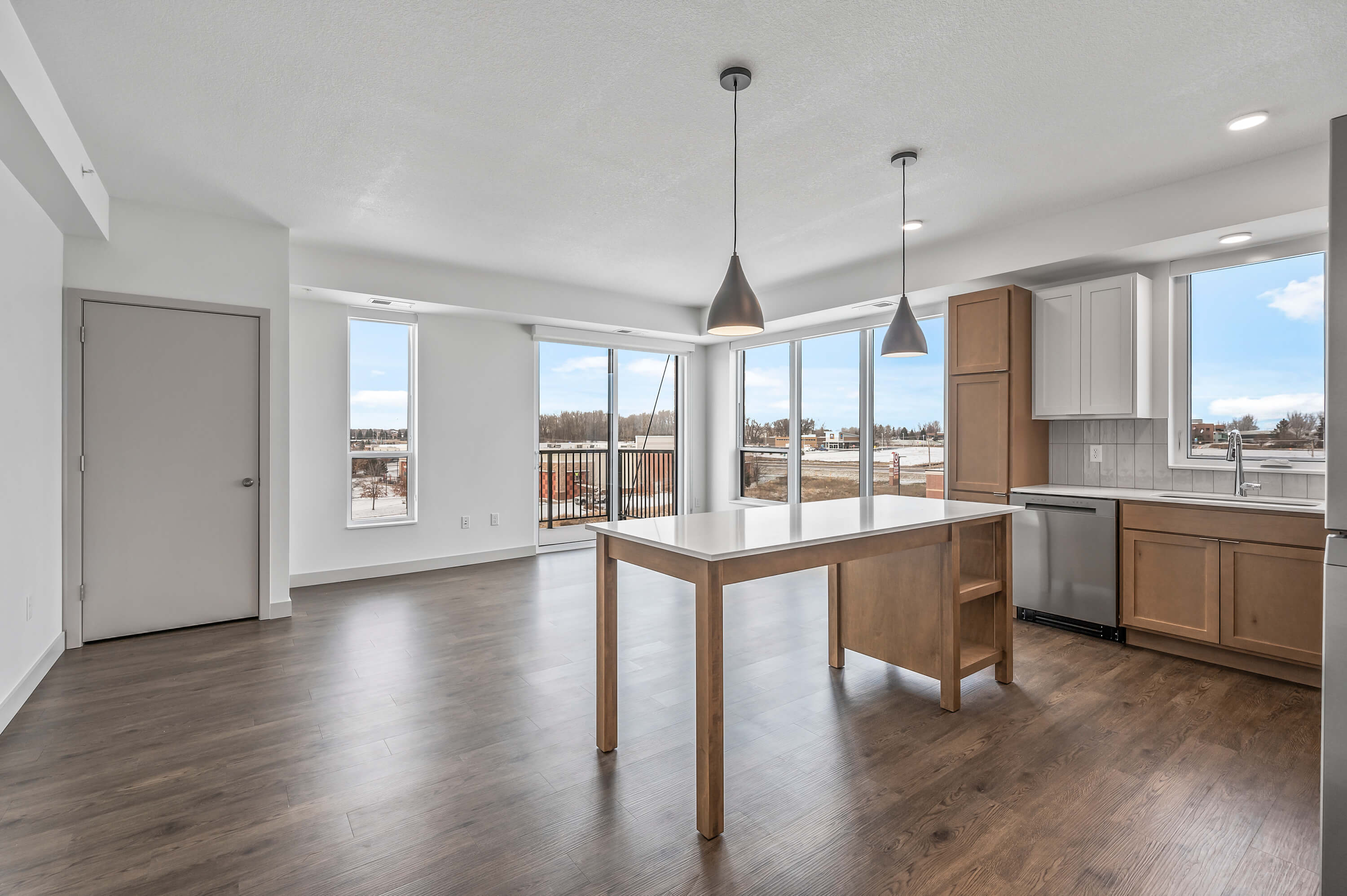 A bright and open kitchen and living area with large windows, pendant lights, and wood flooring.