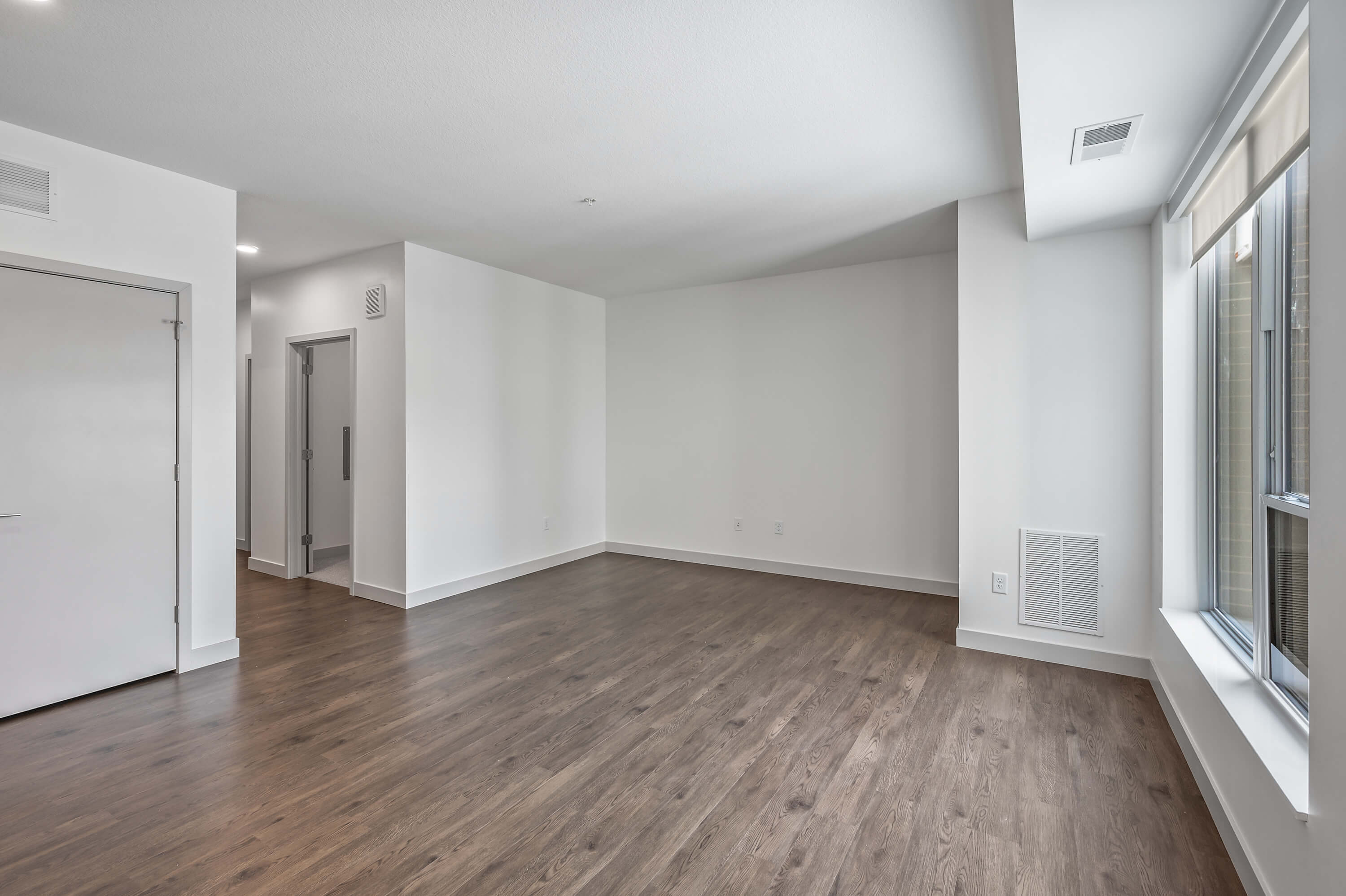 Empty living area with wood floors and large windows, featuring clean white walls.