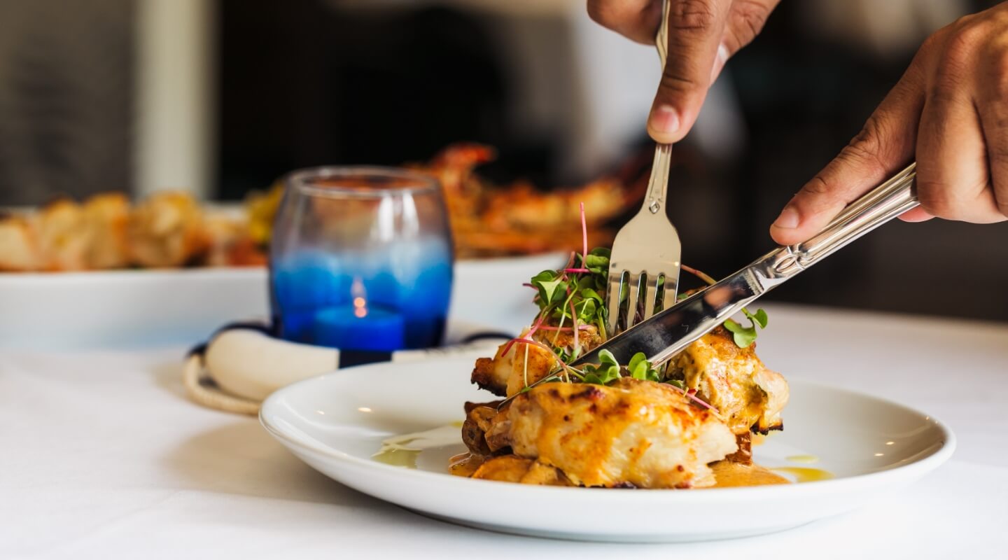 Close-up of a person dining, cutting into a succulent roasted chicken dish garnished with sprouts, with a candlelit ambiance in the background.