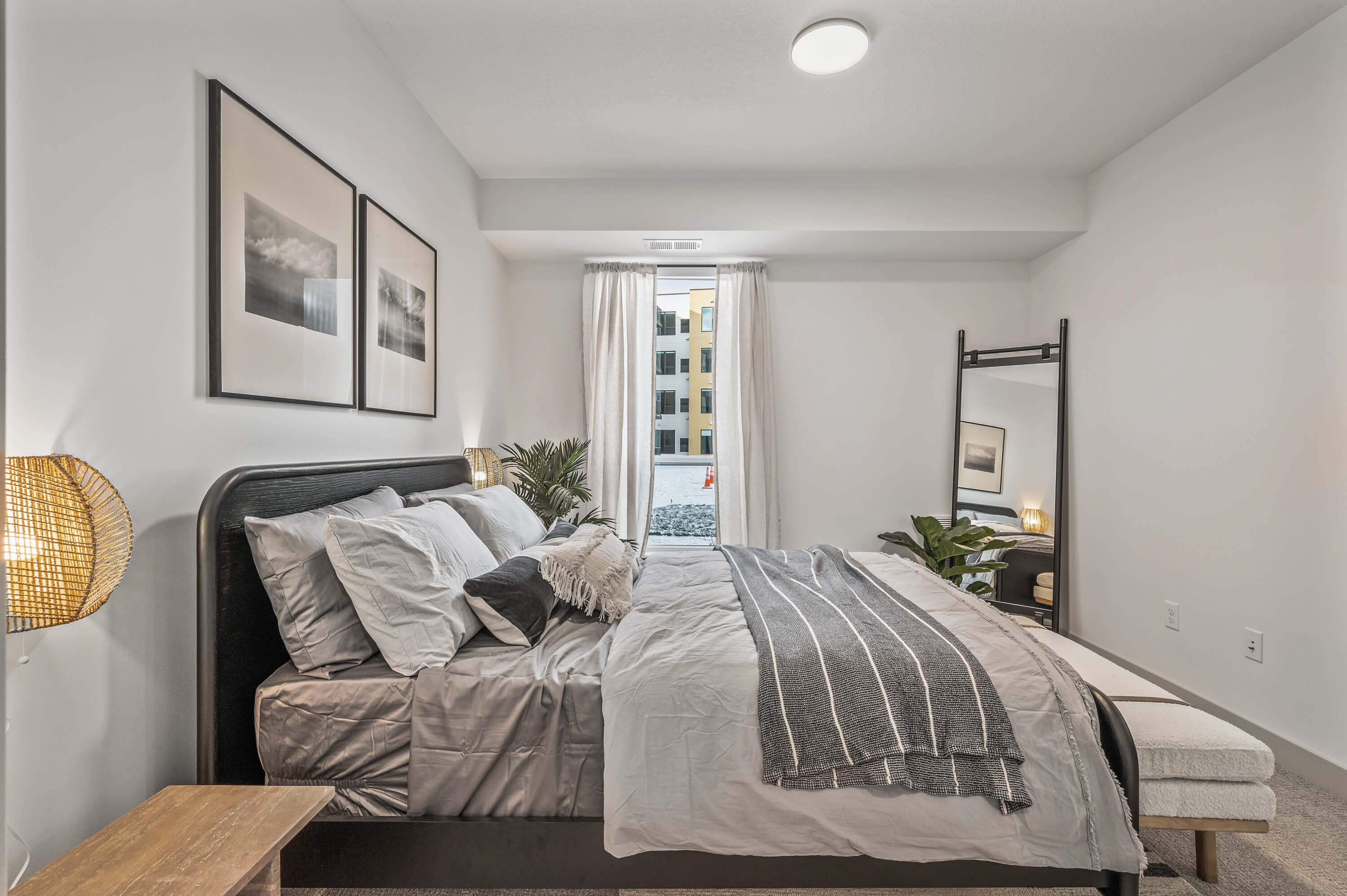 Modern bedroom with a cozy bed, striped blanket, wicker lamps, and a large window with a view of neighboring apartments.