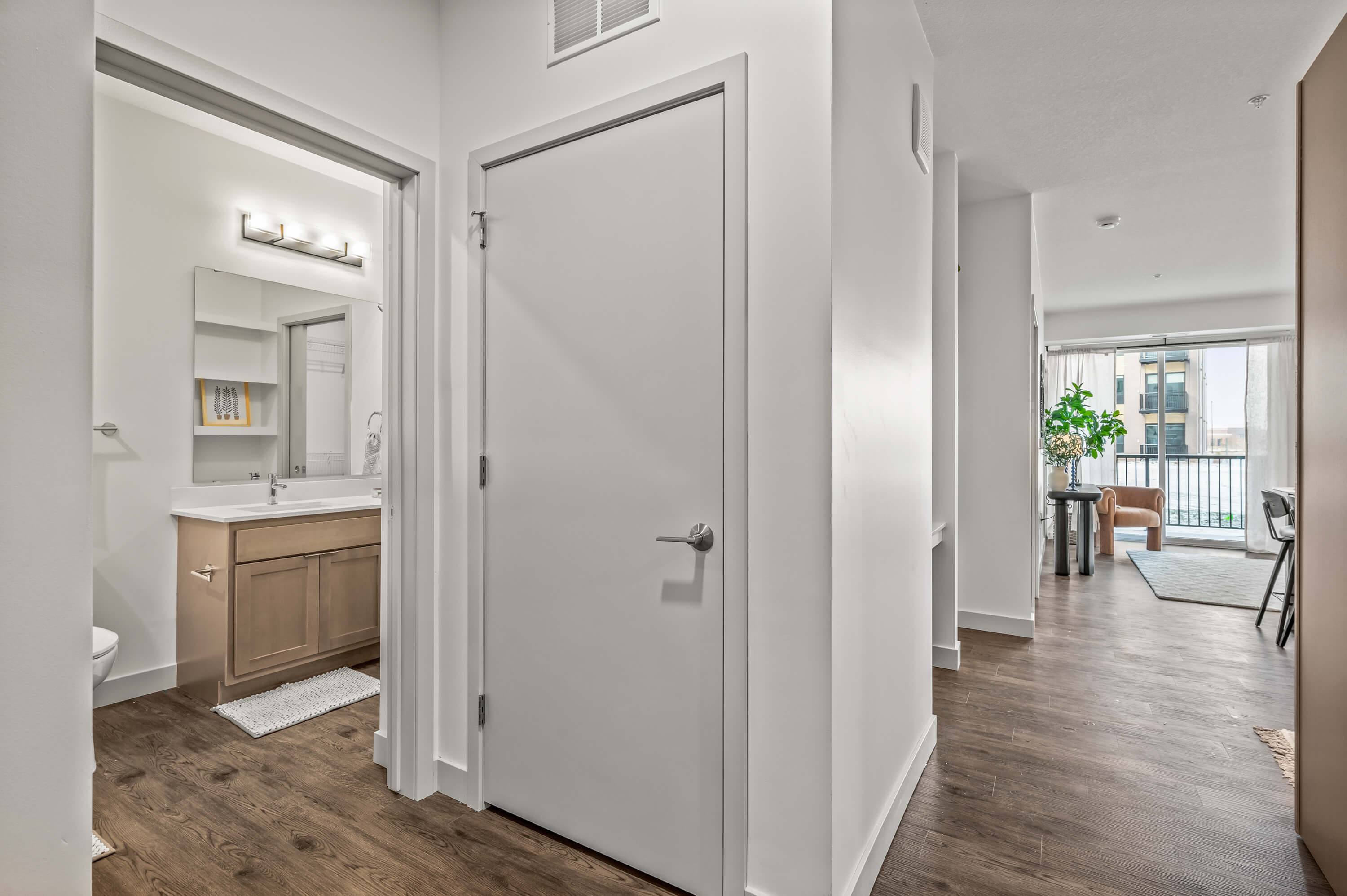 Hallway view featuring a modern bathroom with wooden cabinetry and a vanity mirror, leading into a brightly lit living area with a balcony.