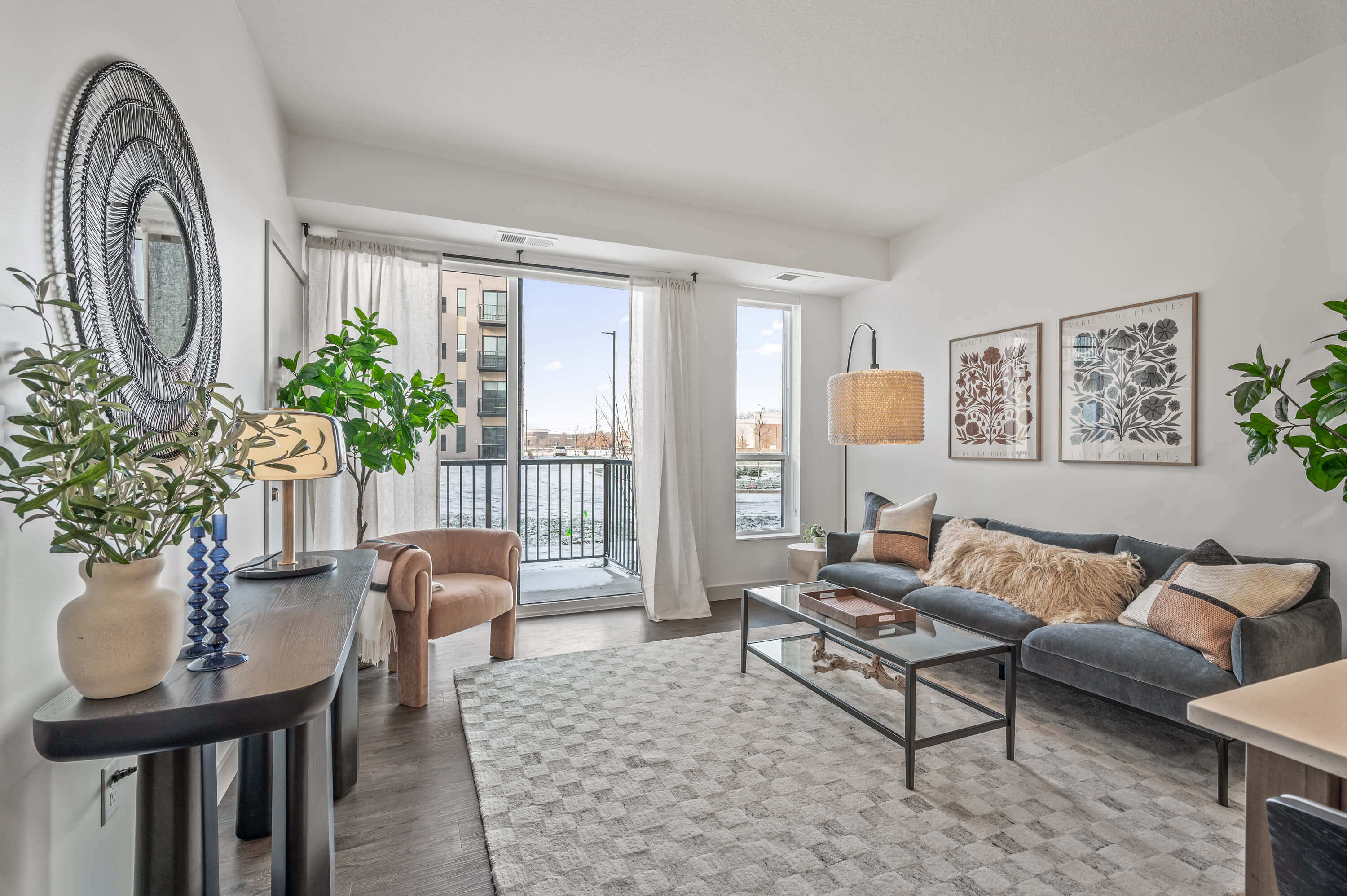 Stylish living room with modern furniture, large windows, and a balcony, featuring neutral tones and greenery accents.