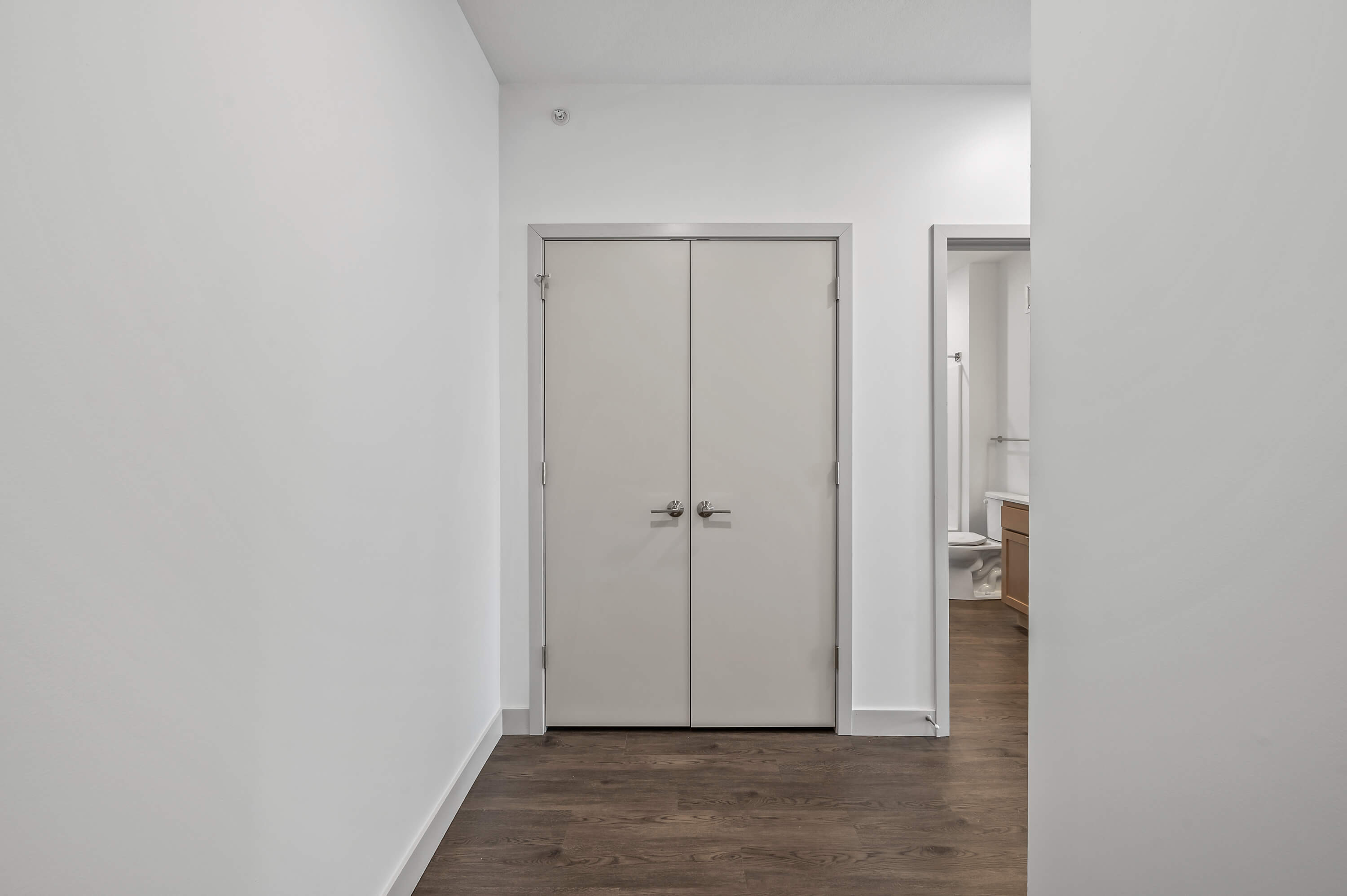 A closet with double doors and adjacent bathroom access, located in a white-painted hallway.