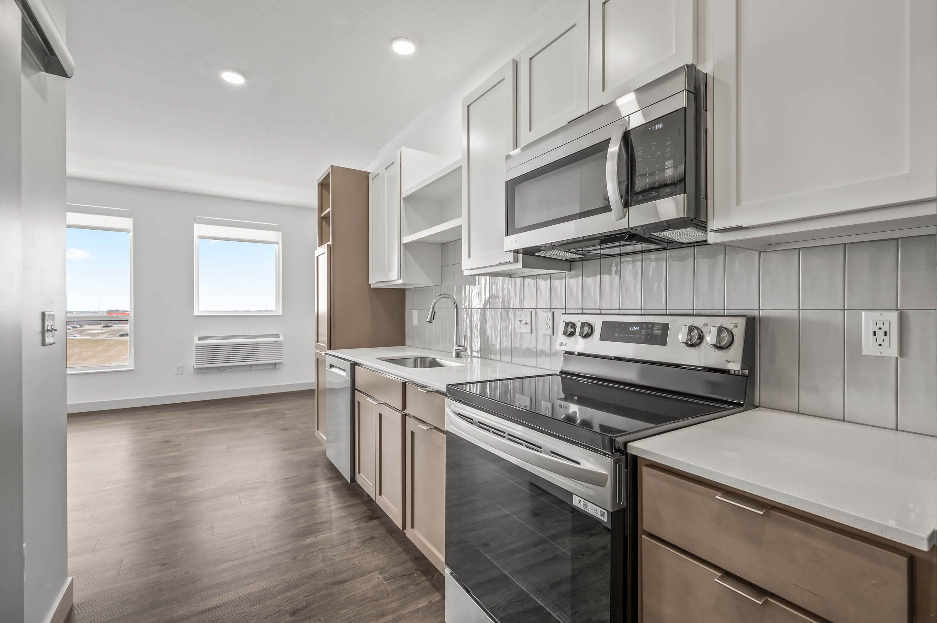 Kitchen with modern stainless steel appliances, white cabinetry, and wood floors, with two large windows in the background.