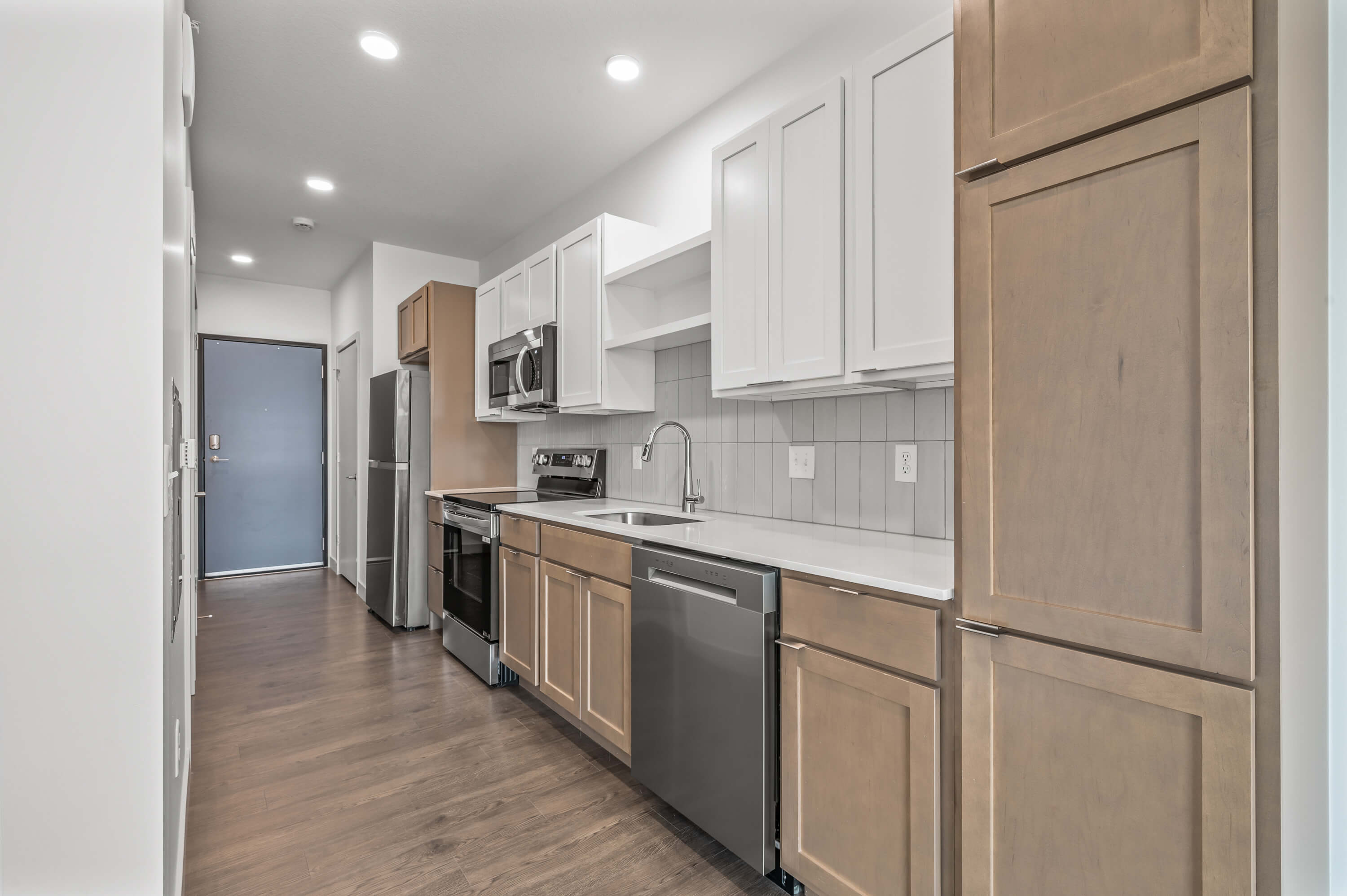 A galley-style kitchen with modern appliances, wooden lower cabinets, white upper cabinets, and a door at the far end.