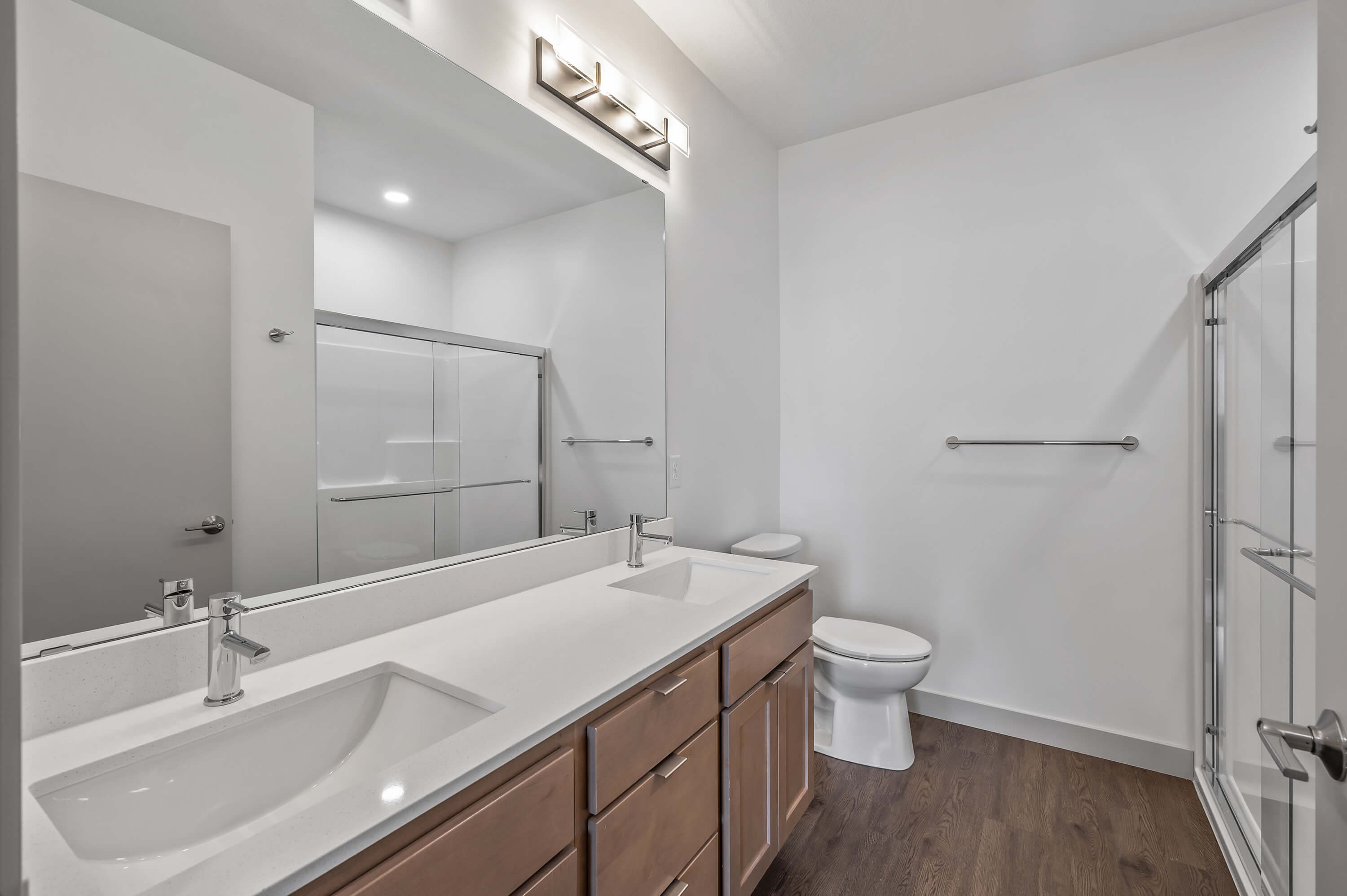 A bathroom featuring a double vanity with white countertops, wooden cabinetry, and a glass-enclosed shower.