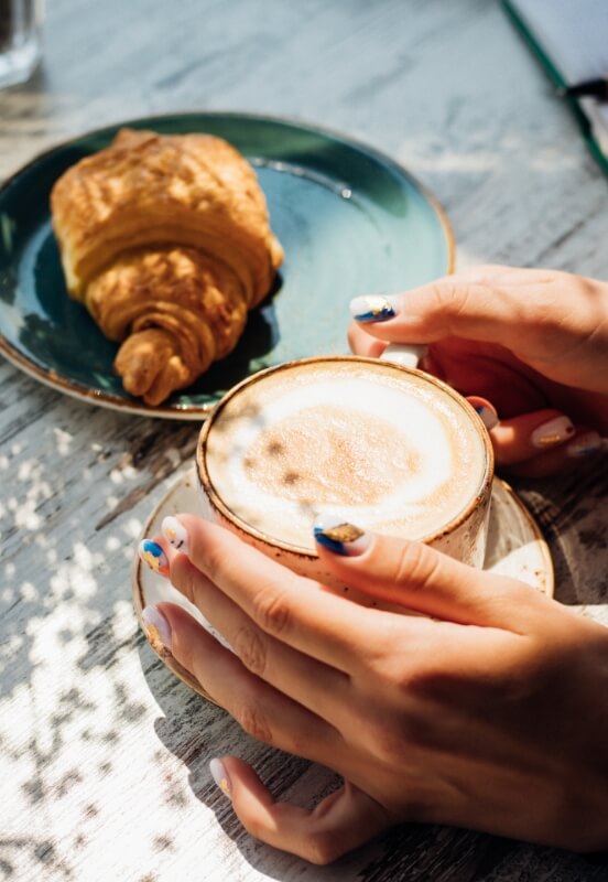 A person enjoying a cappuccino in a ceramic cup, with a fresh croissant on a plate in the background, on a sunlit wooden table.