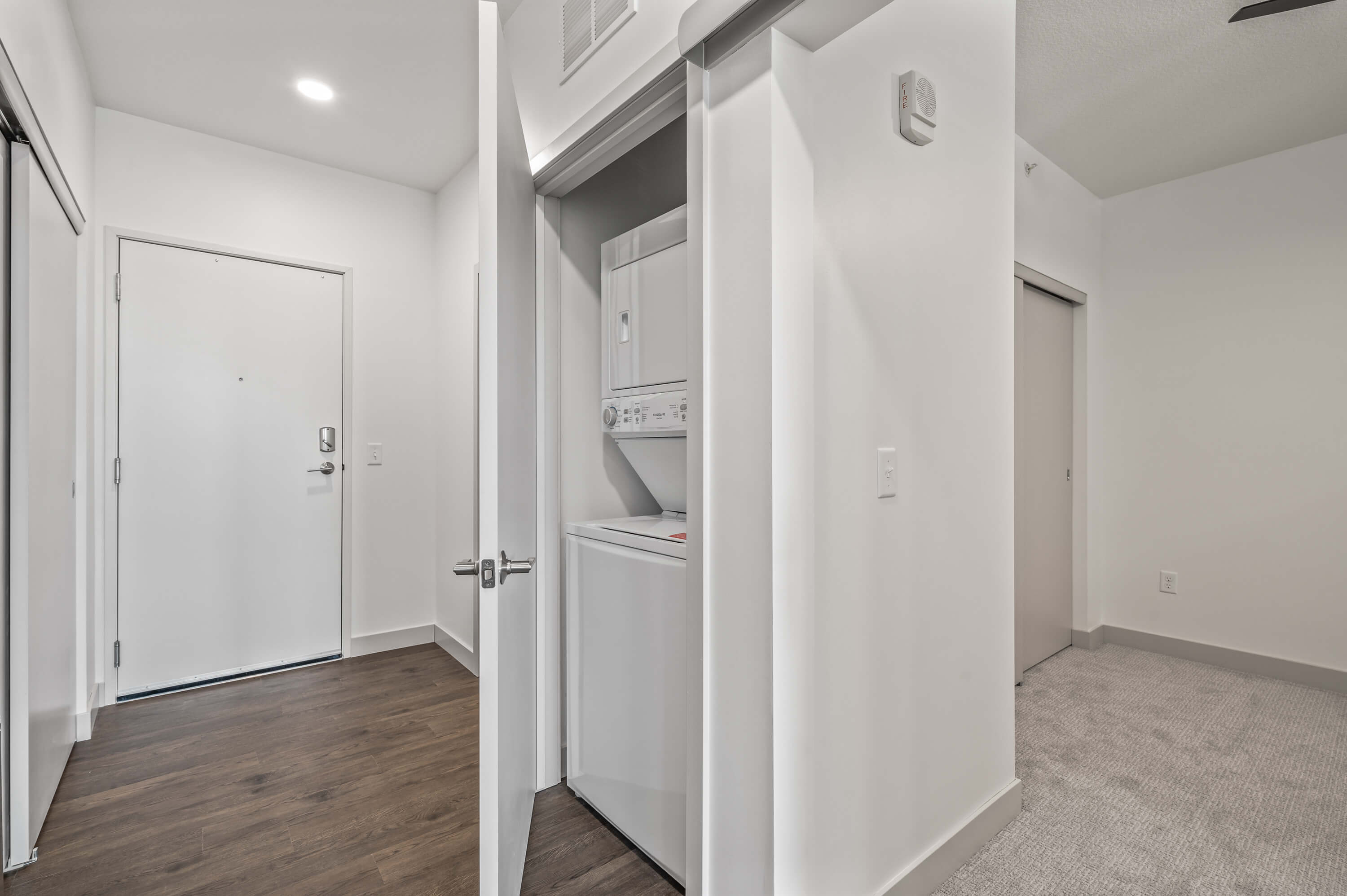 Small laundry area with a stacked washer and dryer unit behind a partially open door, next to a carpeted bedroom.