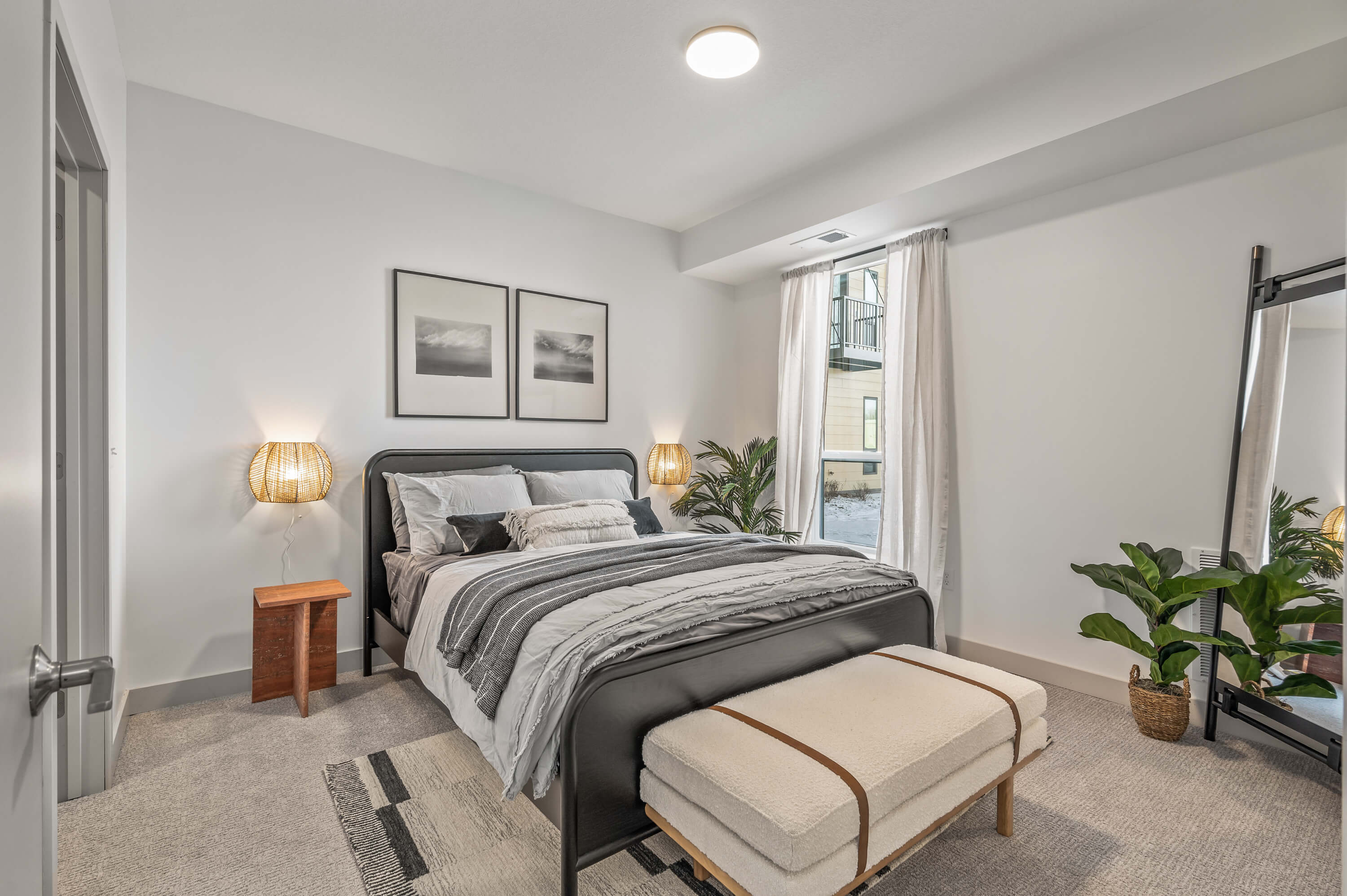 Contemporary bedroom with a cozy bed, natural light from large windows, and minimalist decor including framed artwork and plants.