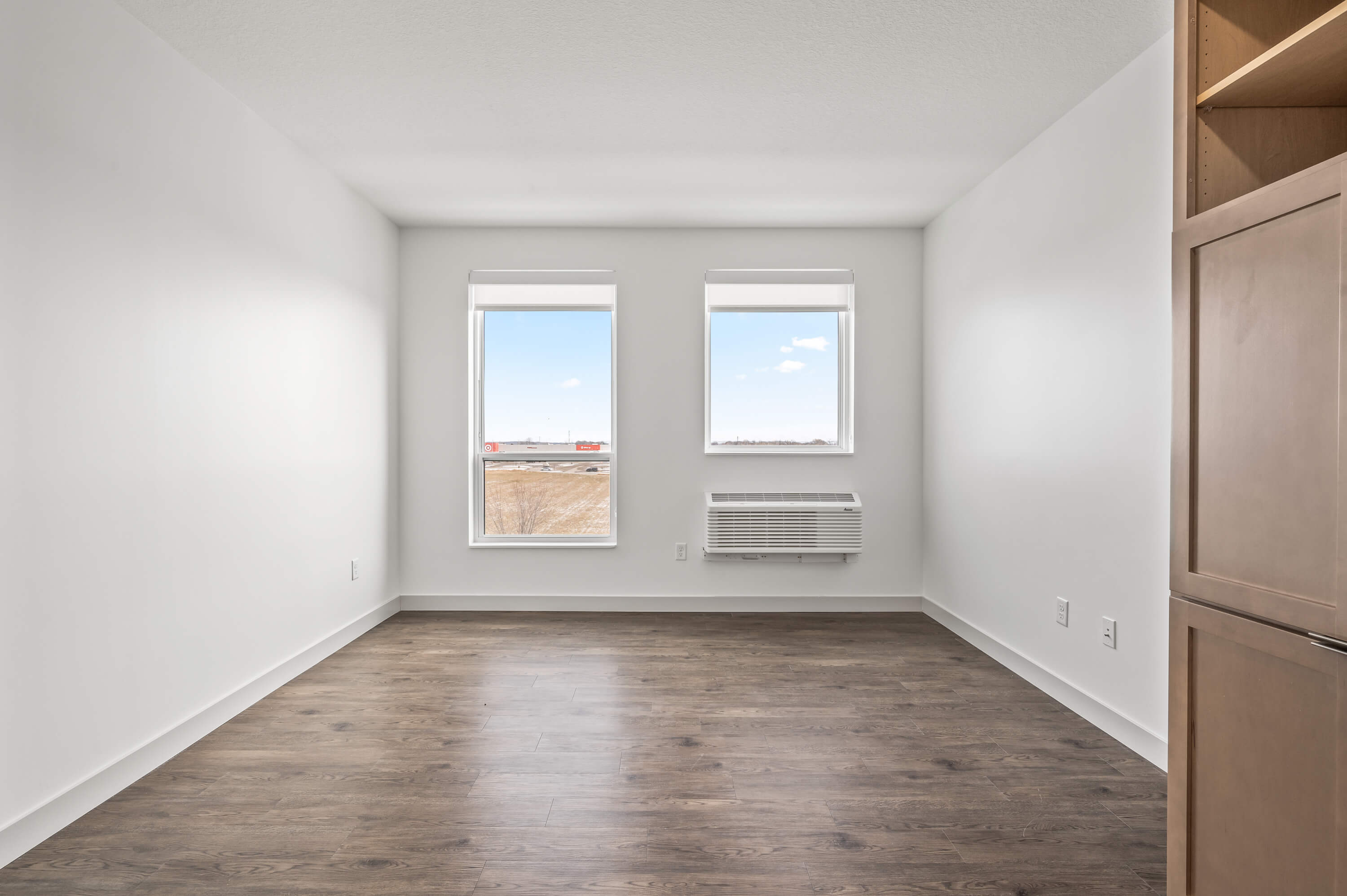 Living area with large windows overlooking an exterior view, air conditioning unit beneath the windows, and wood flooring.