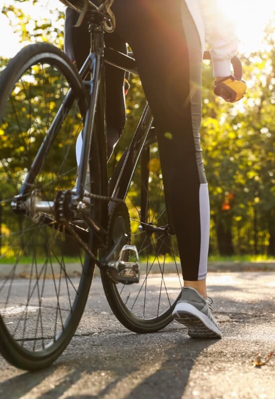 Close-up of a person on a bicycle, focusing on the lower half of their body, with the backdrop of a sunlit tree-lined path.