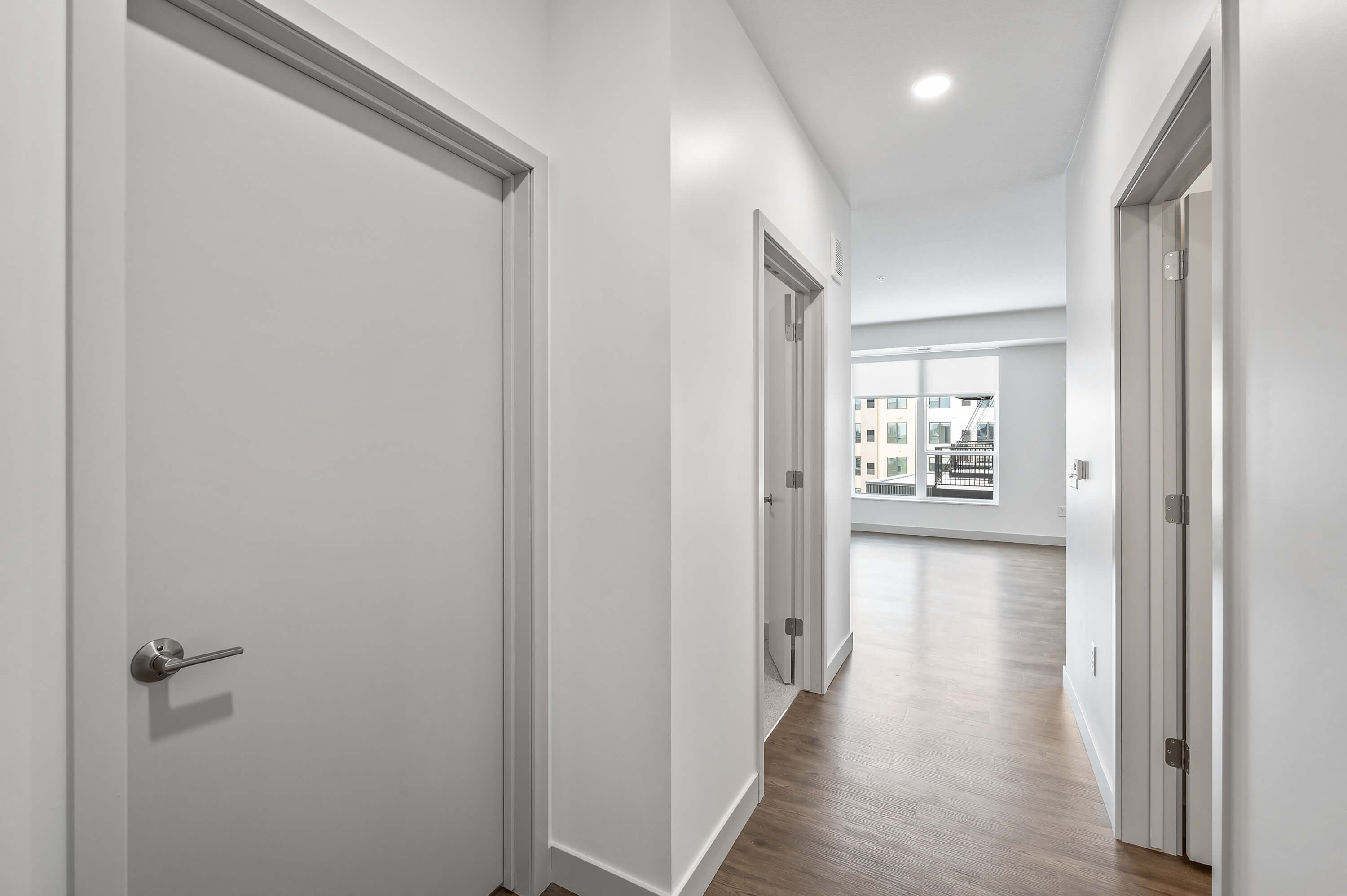 Hallway with white doors and a view into a brightly lit room with large windows.