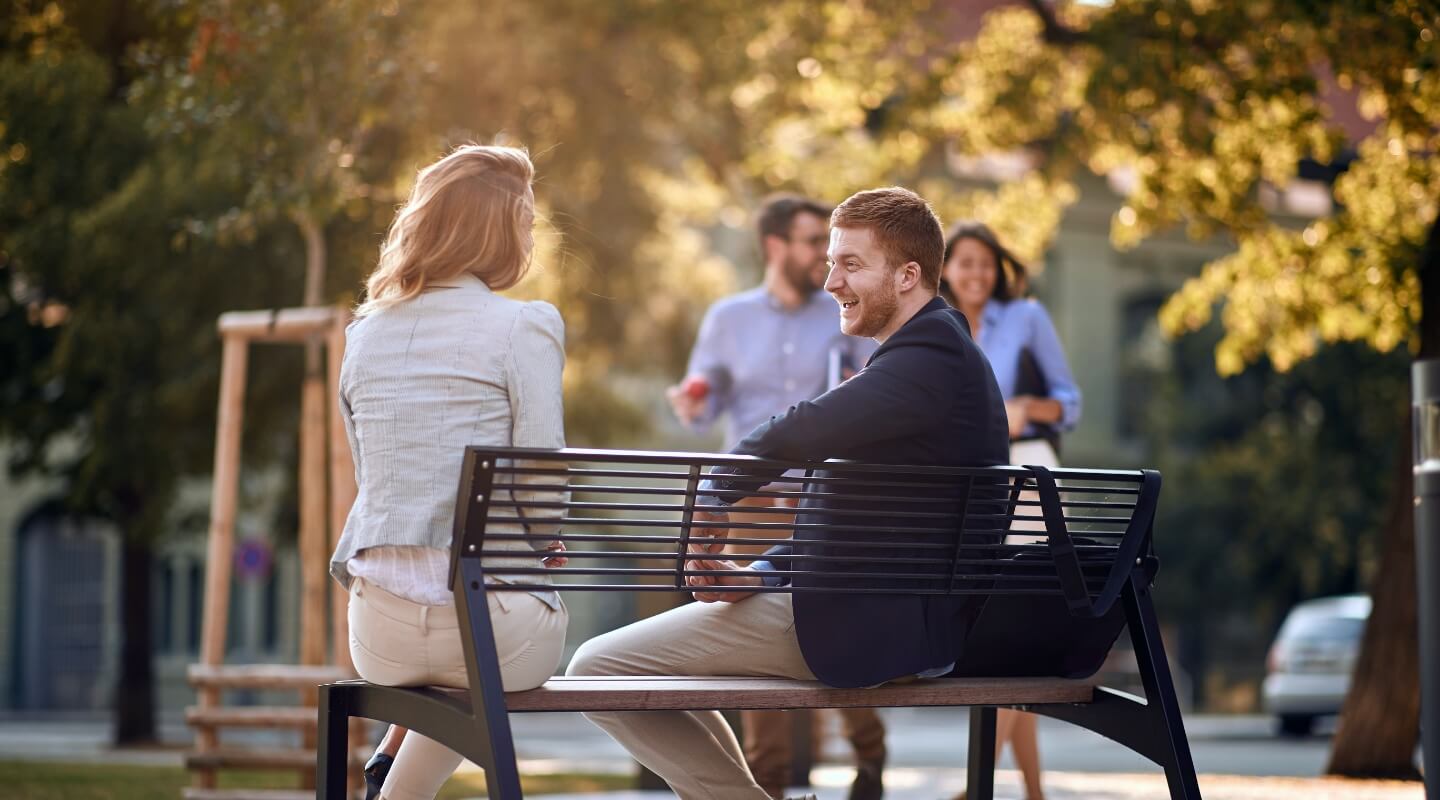 Two people smiling and conversing on a park bench with others walking in the soft-focus background on a sunny day.