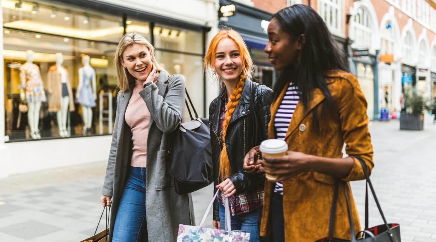 Three friends enjoying a shopping day out, walking and laughing together on a city street with shops in the background.