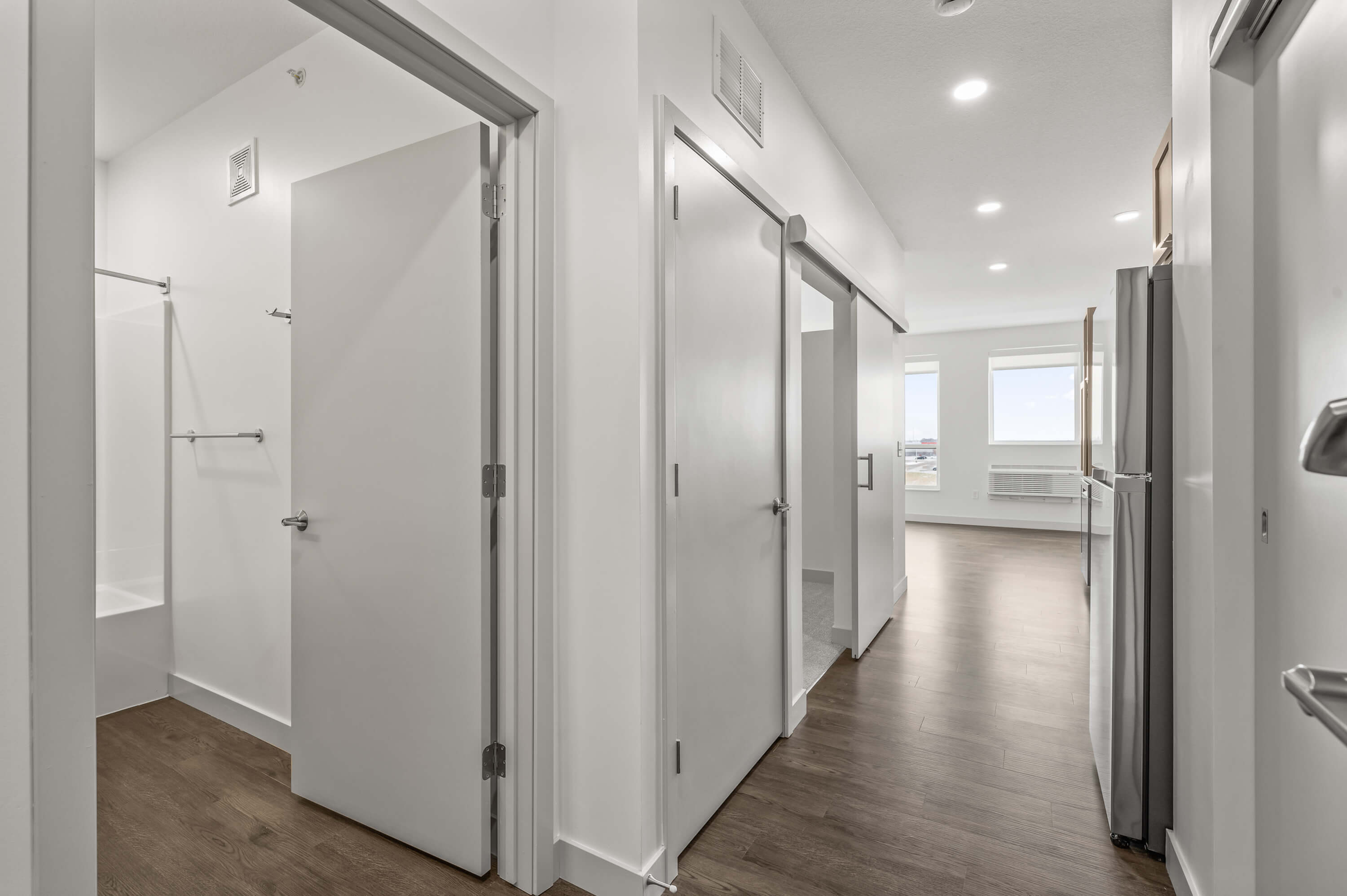 Hallway with doors leading to a bathroom, bedroom, and living area, featuring wood flooring and bright white walls.