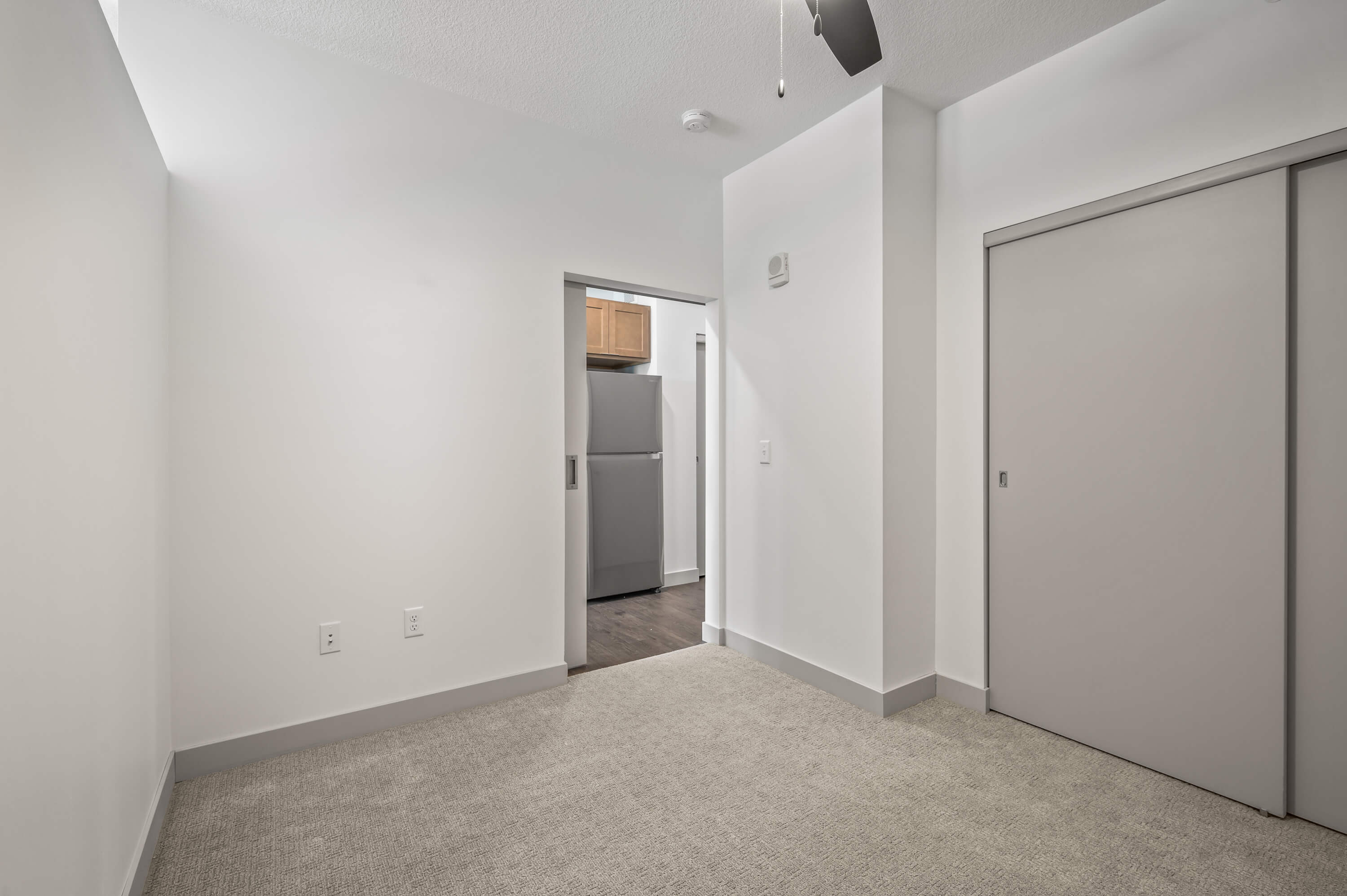 Spacious bedroom with white walls, gray carpet, and an open doorway leading to the kitchen.