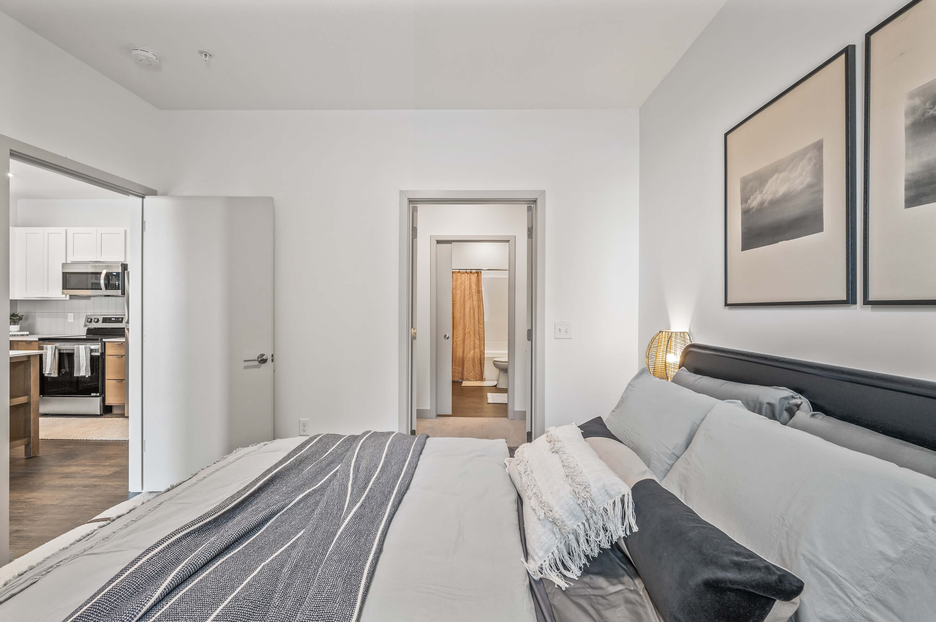 Bedroom view featuring a bed with layered bedding, leading to a bathroom and open kitchen in the background.